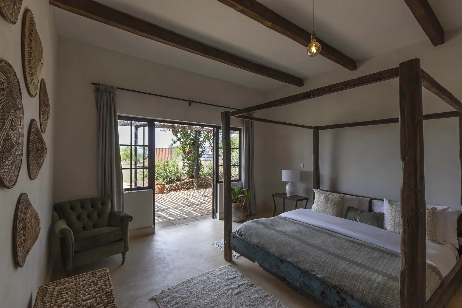 King room one at Lolldaiga Nest, a four poster bed with woven baskets on the wall, a green armchair and doors open to a terrace
