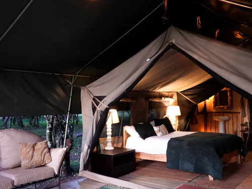 The interior of a canvas safari tent at Maasai Mara Nest, with a bed and lamps under the awning